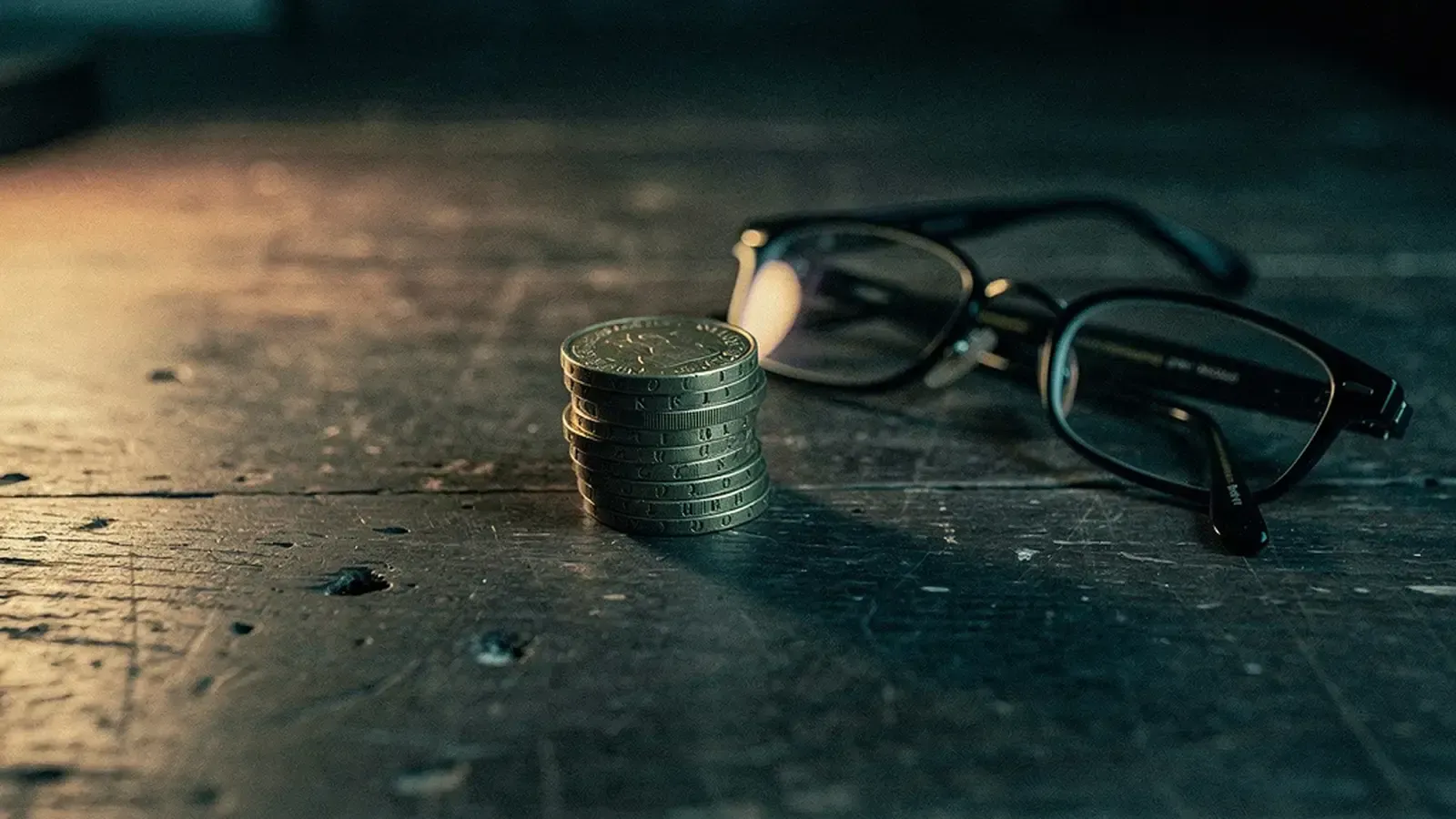 Pound coins and reading glasses on a dark wooden desk in warm lamplight